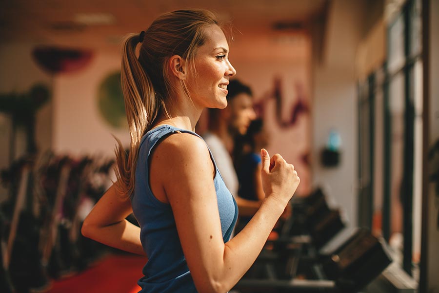 Employee Benefits - Woman Wearing a Blue Tank Top Running on a Treadmill in a Gym with Soft Orange Lighting
