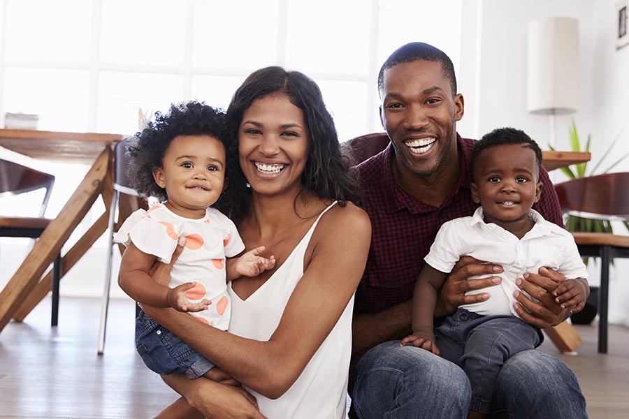 Personal Insurance - Happy Family Sits in Their Home, Kitchen Table Behind Them, Parents Holding Their Baby Daughter and Toddler Son